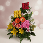 Arreglo floral vertical con rosas rojas, rosas amarillas y rosas rosadas, gerberas naranjas y rosadas, lirios amarillos y blancos sobre fondo rosa desenfocado