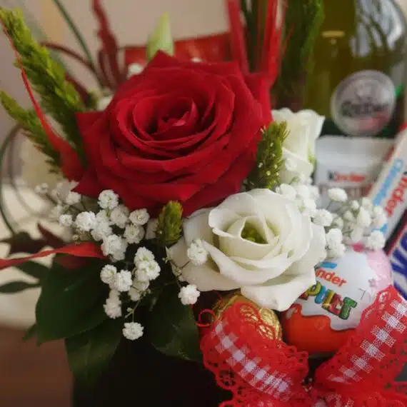 Primer plano extremo de un arreglo floral CARLO con rosas rojas y blancas, paniculata, vino y chocolates Kinder, con gotas de