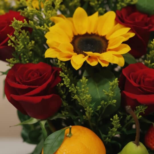 Primer plano extremo del Arreglo Floral Adriano con rosas rojas, girasol central, follaje verde y frutas frescas como naranja