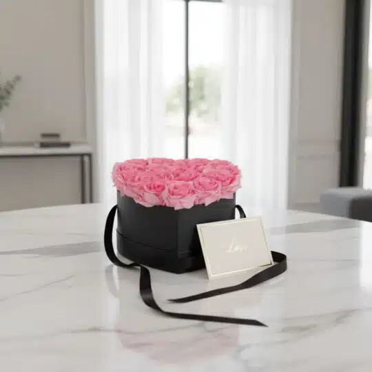 Arreglo Floral Perla con rosas rosadas en caja corazón negra, tarjeta de regalo 'Love' y cinta satinada sobre mesa de mármol.