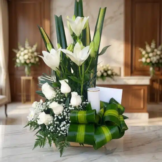 Arreglo Fúnebre Claridad: Lirios y Rosas Blancas en Sala Premium Arreglo fúnebre Claridad con lirios y rosas blancas, vela, follaje verde, en primer plano con fondo de sala de velación premi