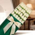 Caja de 24 rosas blancas frescas y elegantes, envueltas en papel verde oscuro con un moño de satén color crema, sobre un fond