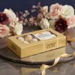 Caja de 8 chocolates Ferrero Rocher dorados sobre una bandeja de mármol con un lazo dorado y un arreglo floral elegante de fo