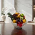 Canasta de frutas gourmet con piña, uvas, manzanas y naranjas, sobre una mesa de madera pulida en un entorno elegante.