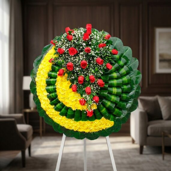 Corona Fúnebre Adela: Elegancia y Respeto en Rosas Rojas Corona fúnebre Adela con rosas rojas y flores blancas sobre base de crisantemos amarillos y hojas verdes, en sala de velación
