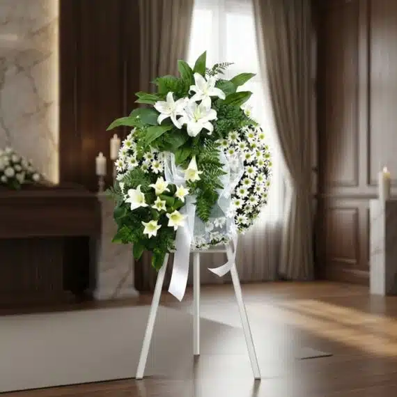 Corona de Homenaje "El Vuelo de Uriel" con Lirios y Margaritas Blancas Corona de homenaje "El Vuelo de Uriel" con lirios blancos, margaritas y follaje verde, sobre un trípode blanco en una sala de