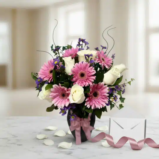 Jarrón Primavera con gerberas rosas, rosas blancas y follaje, sobre mármol con tarjeta de regalo y cinta satinada, listo para