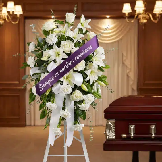 Pedestal conmemorativo de flores blancas, lirios y rosas, con cinta morada que dice 'Siempre en nuestros corazones', junto a