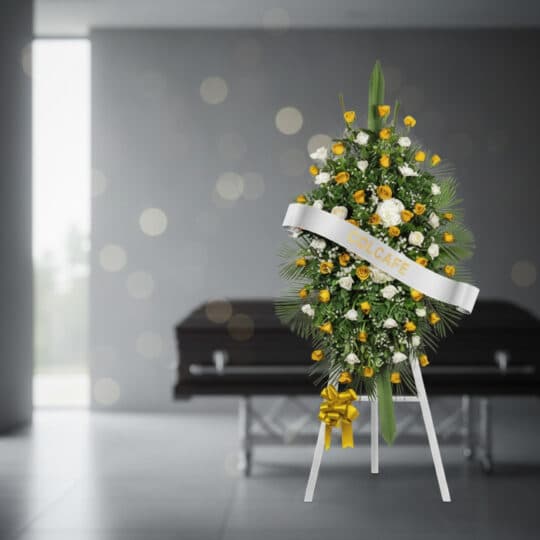 Pedestal fúnebre con rosas amarillas y blancas, follaje verde, y cinta blanca con el nombre 'COLCAFE', en una sala de velació