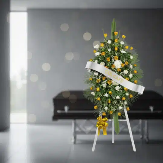 Pedestal fúnebre con rosas amarillas y blancas, follaje verde, y cinta blanca con el nombre 'COLCAFE', en una sala de velació